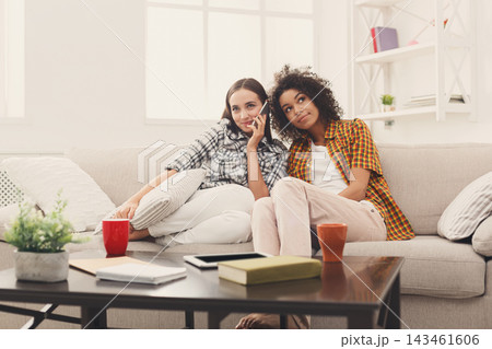Two friends sit on a couch in a living room decorated with plants and books. They engage in conversation while holding drinks. Sunlight enters through the windows. Two friends sit on a couch in a living room decorated with plants and books. They engage in conversation while holding drinks. Sunlight enters through the windows. 143461606
