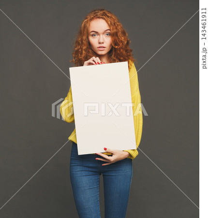 A person stands in a studio holding a blank canvas. The setting has a simple background. The individual looks directly at the camera while dressed in casual clothing. A person stands in a studio holding a blank canvas. The setting has a simple background. The individual looks directly at the camera while dressed in casual clothing. 143461911
