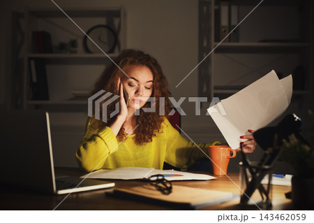 A young woman works late into the night at her desk. She is talking on the phone while holding papers in her hand. A laptop and a cup are nearby. The room is dimly lit with shelves in the background. A young woman works late into the night at her desk. She is talking on the phone while holding papers in her hand. A laptop and a cup are nearby. The room is dimly lit with shelves in the background. 143462059