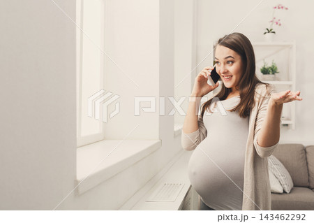 A pregnant woman stands near a window in a living room. She smiles while talking on her phone. Her hand gestures show excitement. A couch and plants are in the background. A pregnant woman stands near a window in a living room. She smiles while talking on her phone. Her hand gestures show excitement. A couch and plants are in the background. 143462292