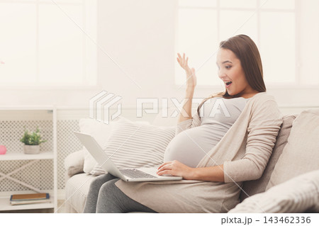 A pregnant woman sits on a light couch with a laptop in front of her. She looks surprised and happy as she raises her hand to express her excitement. Sunlight comes through the windows. A pregnant woman sits on a light couch with a laptop in front of her. She looks surprised and happy as she raises her hand to express her excitement. Sunlight comes through the windows. 143462336