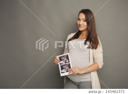 A pregnant woman stands with a smile while holding ultrasound images. She is wearing a simple outfit. The background is plain and neutral. The setting focuses on her and the ultrasound images. A pregnant woman stands with a smile while holding ultrasound images. She is wearing a simple outfit. The background is plain and neutral. The setting focuses on her and the ultrasound images. 143462371