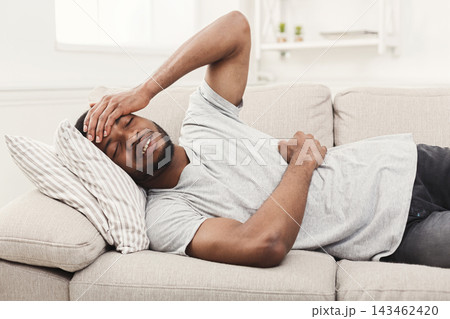 A man lies on a couch in a living room, holding his head with one hand and his stomach with the other. The setting has bright light coming in through a window. 143462420