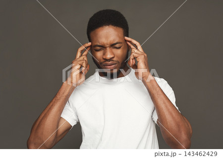 A man stands in a simple indoor location, touching his temples with both hands. He has his eyes closed, showing a thoughtful expression. The background is plain and neutral. 143462429