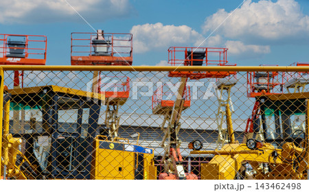 Construction equipment fleet stored behind a yellow mesh fence under blue sky, aerial work platforms and heavy machinery waiting at an outdoor rental yard for industrial projects and maintenance. 143462498