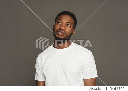 A man stands in a dark area wearing a white shirt. He looks upward with a thoughtful expression. The background is plain and simple. His pose suggests contemplation or curiosity. A man stands in a dark area wearing a white shirt. He looks upward with a thoughtful expression. The background is plain and simple. His pose suggests contemplation or curiosity. 143462522