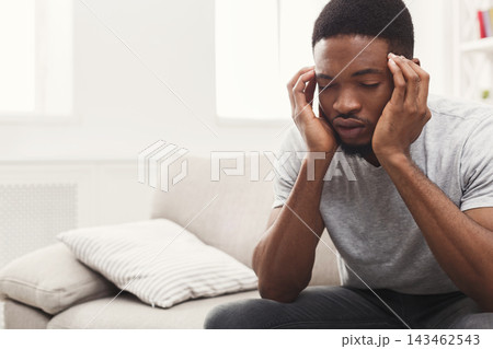 A man sits on a sofa with his hands on his head, appearing deep in thought. He looks down and is surrounded by bright light coming from the window. The room is simple and tidy. 143462543