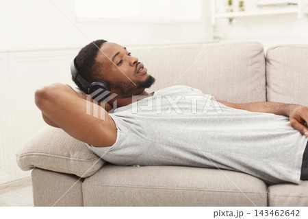 A man is lying on a couch with headphones on, enjoying music. He seems focused and relaxed. Sunlight enters the room, creating a casual atmosphere for resting. 143462642