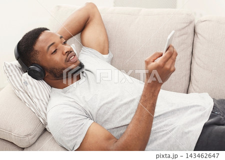 A person rests on a couch with headphones on, looking at a phone. The setting is indoors with soft furnishings. This scene captures a casual moment of leisure and enjoyment. A person rests on a couch with headphones on, looking at a phone. The setting is indoors with soft furnishings. This scene captures a casual moment of leisure and enjoyment. 143462647