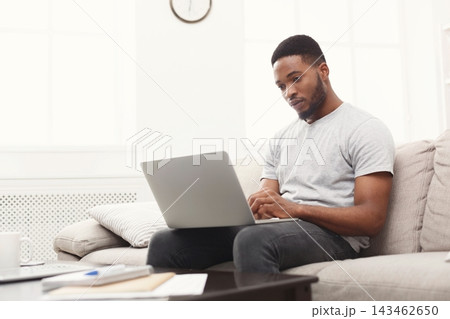 A man is sitting on a couch in a light-filled room. He focuses on his laptop while typing. The setting shows a clock on the wall, giving a sense of home office work during the day. A man is sitting on a couch in a light-filled room. He focuses on his laptop while typing. The setting shows a clock on the wall, giving a sense of home office work during the day. 143462650