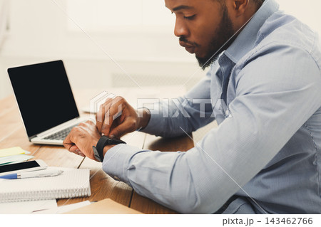 A man sits at a wooden desk looking at his smartwatch. A laptop is open beside him, and there are notepads, pens, and sticky notes on the table. This takes place in a bright office. A man sits at a wooden desk looking at his smartwatch. A laptop is open beside him, and there are notepads, pens, and sticky notes on the table. This takes place in a bright office. 143462766