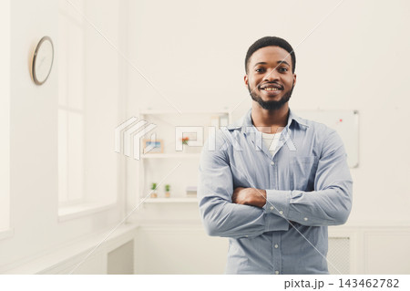A man stands with arms crossed in a bright room. He wears a checkered shirt and smiles. The room has minimal decor, including a clock on the wall. Shelves with small items are visible. A man stands with arms crossed in a bright room. He wears a checkered shirt and smiles. The room has minimal decor, including a clock on the wall. Shelves with small items are visible. 143462782