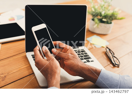 Hands are holding a smartphone while sitting next to a laptop positioned on a wooden table. Surrounding items include a plant, glasses, and colorful notes. The workspace looks organized. Hands are holding a smartphone while sitting next to a laptop positioned on a wooden table. Surrounding items include a plant, glasses, and colorful notes. The workspace looks organized. 143462797