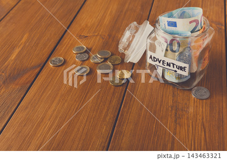A clear jar is filled with banknotes and coins on a wooden table. The jar has a label that says adventure. Some coins are spilled out next to the jar, suggesting saving for future travel. 143463321