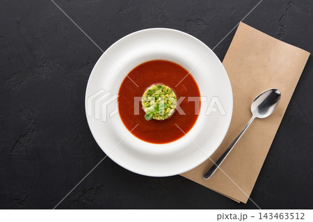A bowl with vibrant red soup sits on a dark surface. Topped with diced green ingredients, the dish is simple. A spoon and a brown napkin are placed beside the bowl. 143463512