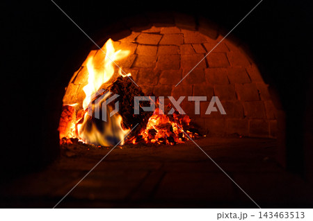 A fire burns in a brick oven, with logs inside catching flames and glowing embers. The light of the fire creates a warm ambiance in the dark setting. Smoke rises gently. 143463513