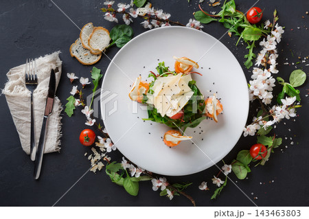 A fresh salad is arranged on a white plate. It features shrimp, cheese slices, and greens. Surrounding the plate are small tomatoes and flowers. Cutlery and bread slices are placed nearby. A fresh salad is arranged on a white plate. It features shrimp, cheese slices, and greens. Surrounding the plate are small tomatoes and flowers. Cutlery and bread slices are placed nearby. 143463803