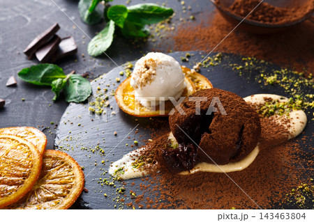 A dark chocolate dessert sits on a black plate with orange slices and a scoop of ice cream. Mint leaves and cocoa powder surround the dish, creating a rich display during dinner. 143463804