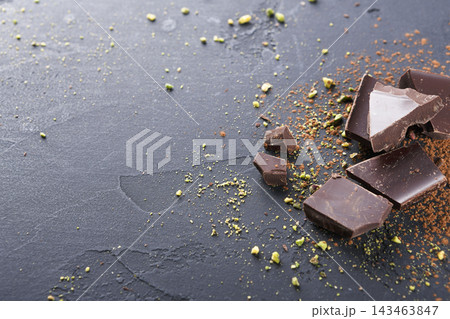 Pieces of chocolate are scattered on a dark surface with crushed nuts around them. This scene captures a moment of preparation in a kitchen setting, providing a glimpse into baking or dessert making. Pieces of chocolate are scattered on a dark surface with crushed nuts around them. This scene captures a moment of preparation in a kitchen setting, providing a glimpse into baking or dessert making. 143463847