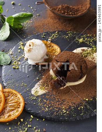 A dessert is served on a dark plate with chocolate cake, a scoop of ice cream, and slices of orange. This scene takes place in a kitchen with a rustic feel. A dessert is served on a dark plate with chocolate cake, a scoop of ice cream, and slices of orange. This scene takes place in a kitchen with a rustic feel. 143463991