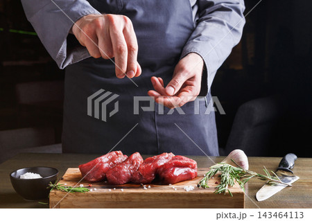 A person sprinkles salt over fresh pieces of meat placed on a wooden cutting board. Fresh herbs are nearby. A knife and bowl of salt are present on the table during cooking time. A person sprinkles salt over fresh pieces of meat placed on a wooden cutting board. Fresh herbs are nearby. A knife and bowl of salt are present on the table during cooking time. 143464113