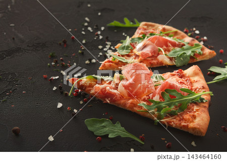 Two slices of pizza sit on a dark surface. The pizza is topped with ham, arugula, and cheese. Sprinkles of salt and pepper are around the slices, adding a touch of flavor. Two slices of pizza sit on a dark surface. The pizza is topped with ham, arugula, and cheese. Sprinkles of salt and pepper are around the slices, adding a touch of flavor. 143464160