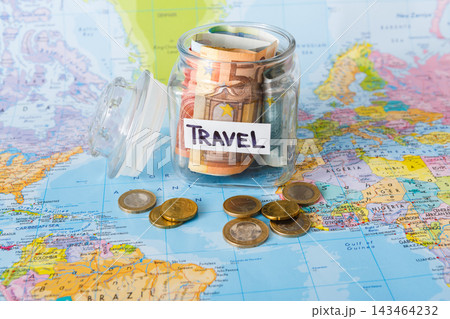 A glass jar contains euro notes labeled travel, sitting on a world map. Several coins lie nearby. This scene suggests planning for a trip and saving money for travel expenses. A glass jar contains euro notes labeled travel, sitting on a world map. Several coins lie nearby. This scene suggests planning for a trip and saving money for travel expenses. 143464232