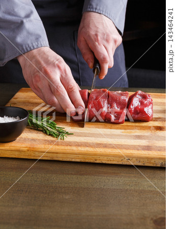 A person slices raw meat into pieces on a wooden cutting board. A bowl of salt and fresh herbs are next to the meat. The scene shows kitchen preparation for a meal. A person slices raw meat into pieces on a wooden cutting board. A bowl of salt and fresh herbs are next to the meat. The scene shows kitchen preparation for a meal. 143464241