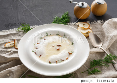 A bowl of creamy soup is placed on a table. The soup is decorated with herbs and spices. Slices of bread are scattered around. Salt and pepper shakers are also on the table. A bowl of creamy soup is placed on a table. The soup is decorated with herbs and spices. Slices of bread are scattered around. Salt and pepper shakers are also on the table. 143464318