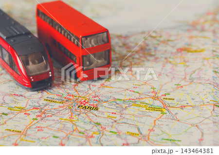 Two toy vehicles, a red bus and a train, sit on a colorful map highlighting Paris. The map shows roads and landmarks in and around the city. Both vehicles are positioned to suggest exploration. Two toy vehicles, a red bus and a train, sit on a colorful map highlighting Paris. The map shows roads and landmarks in and around the city. Both vehicles are positioned to suggest exploration. 143464381