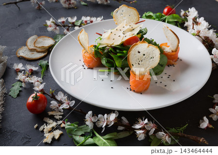 A dish featuring salad greens, salmon slices, and toasted bread shaped like chips is arranged on a white plate. Cherry tomatoes and flowers surround the plate on a dark surface. A dish featuring salad greens, salmon slices, and toasted bread shaped like chips is arranged on a white plate. Cherry tomatoes and flowers surround the plate on a dark surface. 143464444