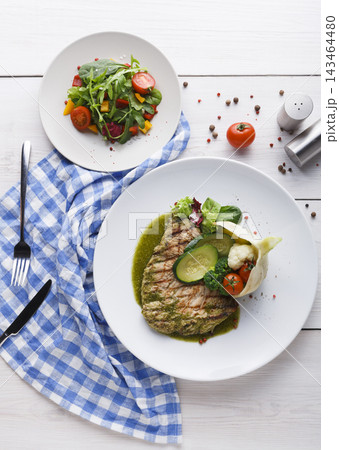 A grilled fish fillet is placed on a white plate with vegetables and sauce. A salad is on a small plate beside it. A blue checkered napkin rests on the table with some seasoning nearby. A grilled fish fillet is placed on a white plate with vegetables and sauce. A salad is on a small plate beside it. A blue checkered napkin rests on the table with some seasoning nearby. 143464480