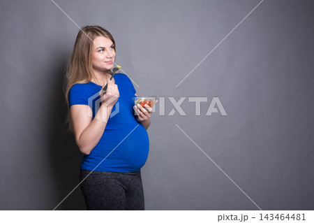 A woman in a blue shirt stands with a bowl of salad in one hand and a fork in the other. She is smiling and looking at the salad. Her belly shows she is pregnant. The setting has a gray background. A woman in a blue shirt stands with a bowl of salad in one hand and a fork in the other. She is smiling and looking at the salad. Her belly shows she is pregnant. The setting has a gray background. 143464481