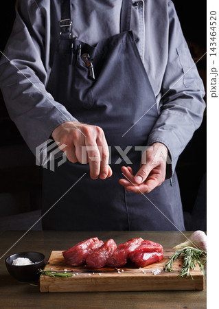 A chef stands at a wooden table and seasons raw meat with salt. The scene is set in a dark kitchen, highlighting the chef's focused activity. Fresh herbs are present nearby. A chef stands at a wooden table and seasons raw meat with salt. The scene is set in a dark kitchen, highlighting the chef's focused activity. Fresh herbs are present nearby. 143464520