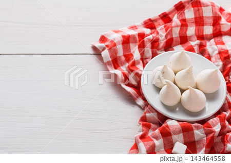 Six meringue cookies are arranged on a white plate. The plate sits on a red checkered cloth. Soft lighting creates a simple display of the cookies. 143464558