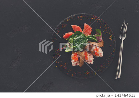 A fish dish is presented on a round black plate with greens and grapefruit slices. Fork and knife sit beside the plate as it awaits to be enjoyed. The setting looks ready for a meal. 143464613