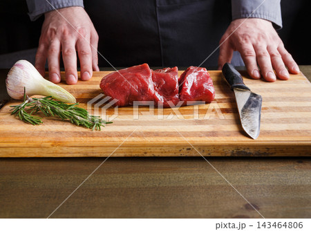 A chef stands with hands on a wooden cutting board. There is a piece of raw meat, fresh herbs, and a knife nearby. The kitchen environment suggests preparation for cooking. 143464806