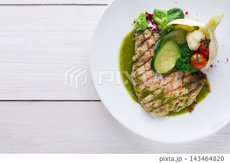 A piece of grilled fish is placed on a white plate. The dish includes vegetables like cucumber, tomatoes, and greens. A green sauce is drizzled around the food. The setting is simple. A piece of grilled fish is placed on a white plate. The dish includes vegetables like cucumber, tomatoes, and greens. A green sauce is drizzled around the food. The setting is simple. 143464820