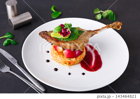 A duck leg is placed on a mound of rice beside a sauce made with fruit. There are greens and garnishes on top. The setting shows a simple dining scene. A duck leg is placed on a mound of rice beside a sauce made with fruit. There are greens and garnishes on top. The setting shows a simple dining scene. 143464821