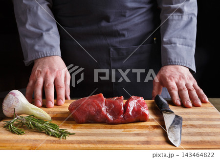A chef stands at a wooden cutting board with a piece of meat, knife, and fresh herbs. The setting is a kitchen where food preparation takes place. A chef stands at a wooden cutting board with a piece of meat, knife, and fresh herbs. The setting is a kitchen where food preparation takes place. 143464822
