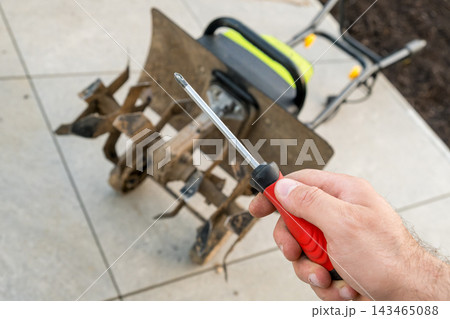 In an outdoor setting, a technician holds a screwdriver for repairing a garden tiller machine. The technician is preparing tools to fix the tillers components on a concrete surface. 143465088