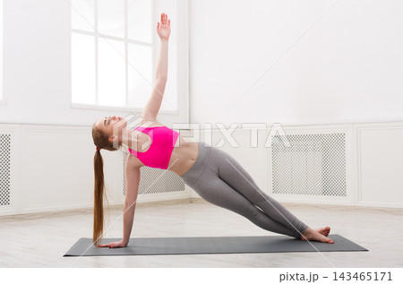 A young woman is performing a side plank yoga pose on a mat in a bright room. She has long hair and wears a pink top and gray pants. Sunshine comes through the windows. A young woman is performing a side plank yoga pose on a mat in a bright room. She has long hair and wears a pink top and gray pants. Sunshine comes through the windows. 143465171