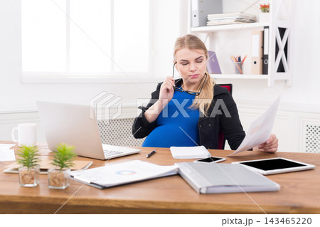 A pregnant woman sits at her desk in a bright office. She holds a pen in one hand and looks at papers. A laptop and tablet are on the desk. Plants are placed nearby. A pregnant woman sits at her desk in a bright office. She holds a pen in one hand and looks at papers. A laptop and tablet are on the desk. Plants are placed nearby. 143465220