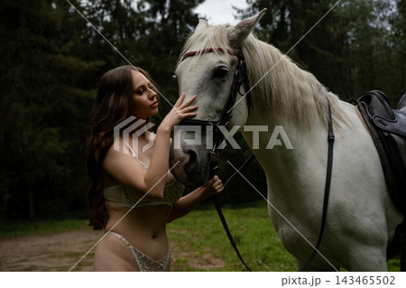 Woman in lace lingerie stroking a white horse in a clearing Woman in lace lingerie stroking a white horse in a clearing 143465502