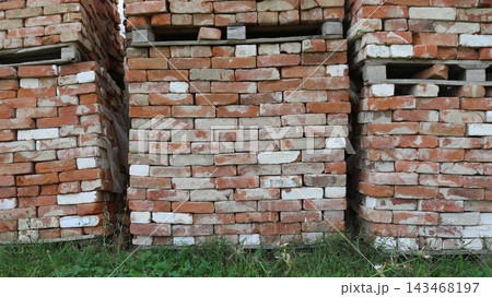 Stacks of old red brick on wooden pallets, neatly arranged in rows outdoors on the green grass. Textured building material with chips and traces of mortar. Stacks of old red brick on wooden pallets, neatly arranged in rows outdoors on the green grass. Textured building material with chips and traces of mortar. 143468197