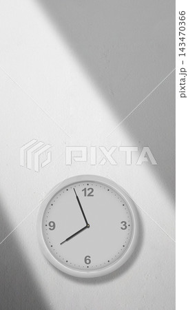 Minimalist white wall clock mounted on a white wall with diagonal sunlight shadows, showing the time. Quotes background 143470366