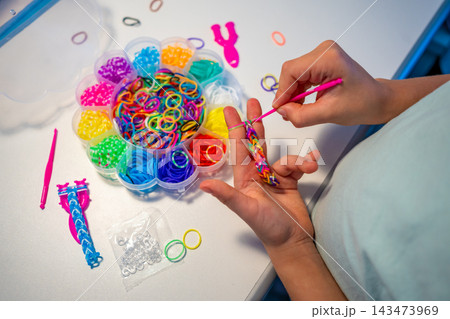 Child weaves a colorful bracelet from rubber bands using fingers and a crochet hook. Creative hobby develops fine motor skills, hand eye coordination and concentration through practice Child weaves a colorful bracelet from rubber bands using fingers and a crochet hook. Creative hobby develops fine motor skills, hand eye coordination and concentration through practice 143473969