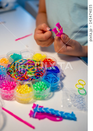 Child weaves a colorful bracelet from rubber bands using fingers and a crochet hook. Creative hobby develops fine motor skills, hand eye coordination and concentration through practice 143474035