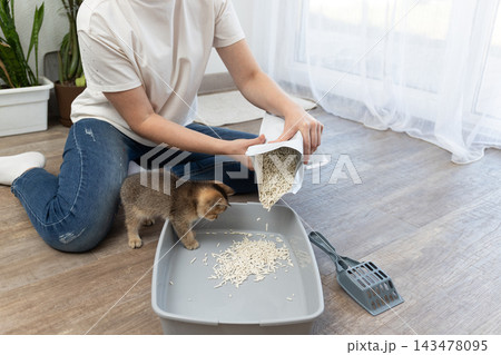 Pouring cat litter into a litter box as a kitten watches nearby in a home scene Pouring cat litter into a litter box as a kitten watches nearby in a home scene 143478095