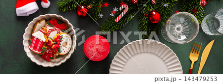 A Christmas table setting featuring an empty plate, napkin and gingerbread ,cutlery and a candle on a deep green background. Spruse branches and red tree decorations create a festive atmosphere A Christmas table setting featuring an empty plate, napkin and gingerbread ,cutlery and a candle on a deep green background. Spruse branches and red tree decorations create a festive atmosphere 143479253